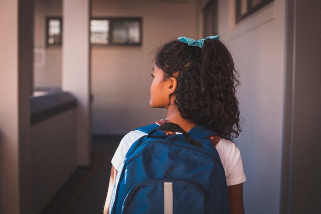 Rear view of mixed race schoolgirl wearing backpack standing in school corridor - Download Free Stock Photos Pikwizard.com