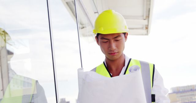 A young Asian male engineer in a safety vest and helmet reviews blueprints on a construction site, with copy space. His focused demeanor suggests he is carefully planning or inspecting work to be done.