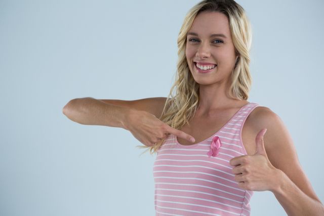 Portrait of smiling woman showing thumbs up while pointing on pink ribbon against gray background
