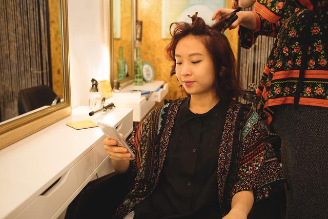 Woman using mobile phone while getting her hair straightened - Download Free Stock Photos Pikwizard.com