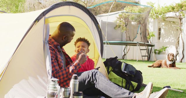 Happy african american father and his son sitting in tent and using tablet in garden. Spending quality time at home.