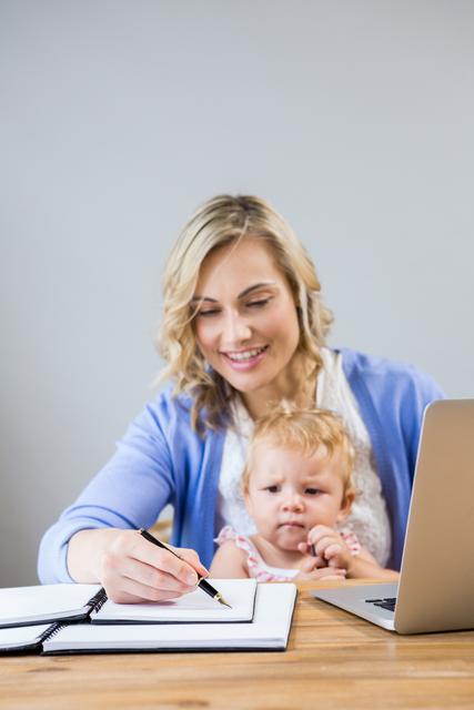 Mother holding baby girl while writing notes in personal organizer - Download Free Stock Photos Pikwizard.com