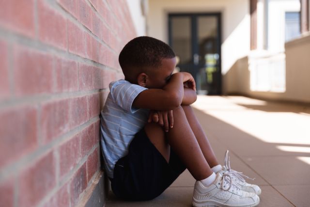 Side view of african american elementary schoolboy hugging knees while sitting on floor in corridor - Download Free Stock Photos Pikwizard.com