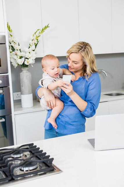 Mother and baby boy using mobile phone in kitchen - Download Free Stock Photos Pikwizard.com