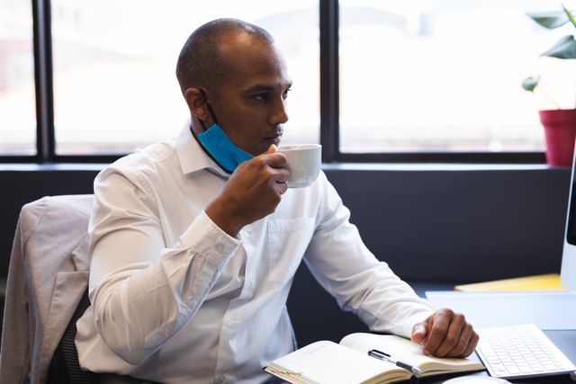 Mixed race businessman wearing lowered face mask drinking coffee sitting at desk. health and hygiene in workplace during coronavirus covid 19 pandemic.