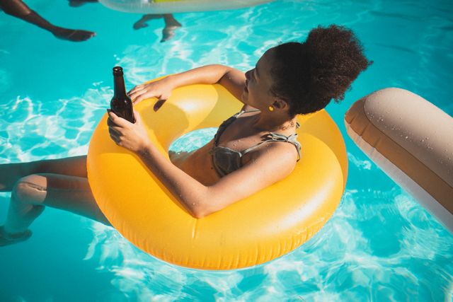 Mixed race woman in inflatable swimming ring in the pool sunbathing, drinking beer and relaxing - Download Free Stock Photos Pikwizard.com