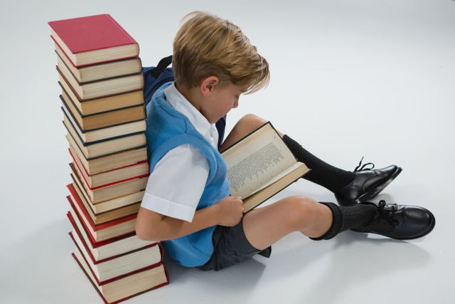 Schoolboy reading book while sitting against books stack - Download Free Stock Photos Pikwizard.com