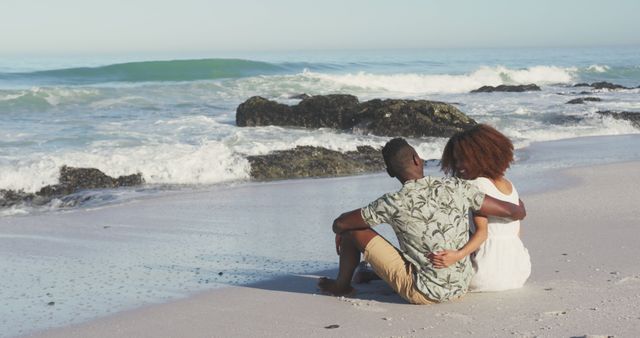 Rear view of happy diverse couple embracing sitting on beach by the sea - Download Free Stock Photos Pikwizard.com