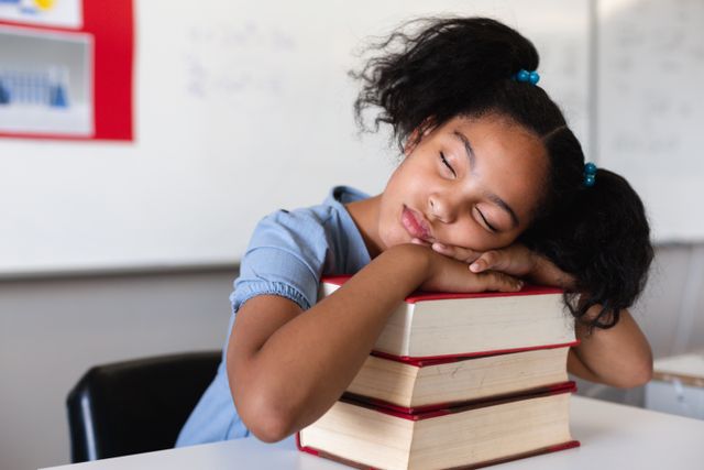 Biracial elementary schoolgirl sleeping on stacked books at desk in classroom. unaltered, education, sleeping, relaxation and school concept.