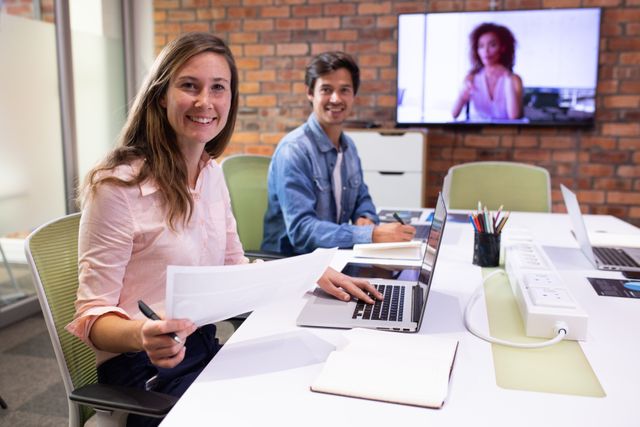 Portrait of a happy Caucasian businesswoman and a businessman working in a modern creative office, having a video conference with a television screen behind them on brick wall, sitting at a desk smiling to camera.