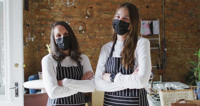 Two caucasian women wearing face masks and aprons, looking at camera. small independent cafe business during coronavirus covid 19 pandemic.