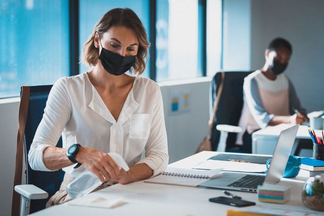 Two diverse businesswomen wearing face masks sitting in office, one disinfecting her desk - Download Free Stock Photos Pikwizard.com