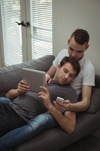 Gay couple using digital tablet and mobile phone while lying on sofa - Download Free Stock Photos Pikwizard.com