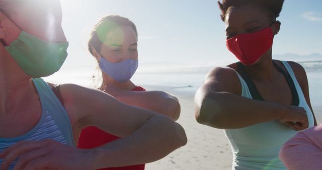 Group of diverse female friends wearing face masks and touching elbows at the beach. healthy active lifestyle, outdoor fitness and wellbeing.