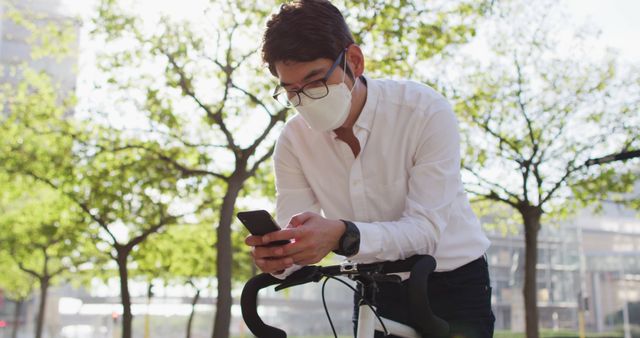 Asian man wearing face mask using smartphone while leaning on his bicycle on the street. health protection and safety during covid-19 pandemic concept