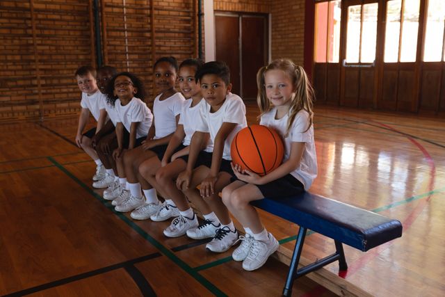Side view of schoolkids with basketball sitting on bench looking at camera in basketball court