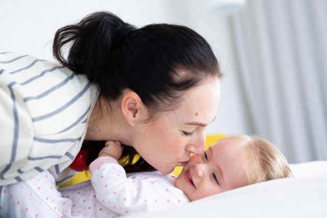 Happy caucasian mother kissing smiling baby daughter lying on bed - Download Free Stock Photos Pikwizard.com