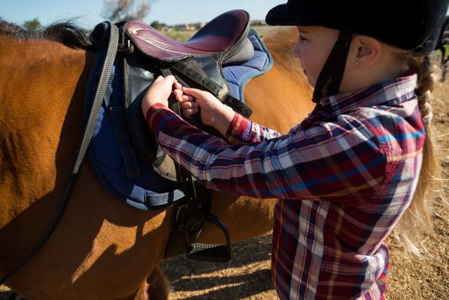 Close-up of girl adjusting saddle on horse in ranch