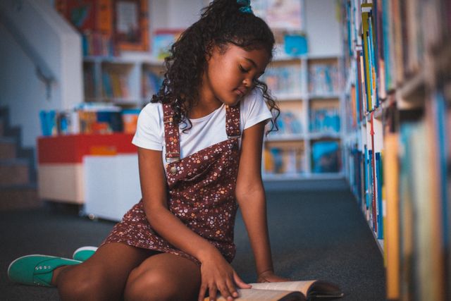 Mixed race schoolgirl sitting on floor reading book in school library - Download Free Stock Photos Pikwizard.com