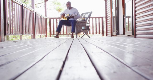 Happy senior african american man spending time in log cabin, playing guitar on balcony. Free time, domestic life and nature concept.