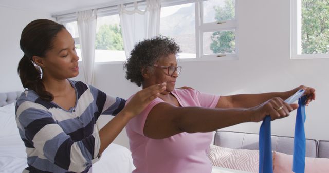 Senior african american woman and female physiotherapist holding resistance band in living room. Medicine, healthcare and lifestyle.
