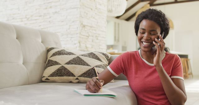 Image of happy african american woman sitting on floor, using smartphone and making notes. home office and online working concept.