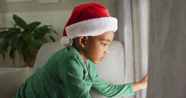 Sad african american boy wearing santa hat looking outside window at christmas time. christmas, festivity and tradition at home.
