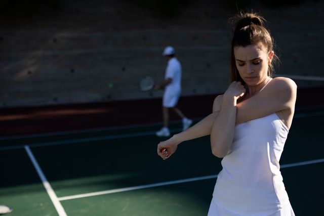 Front view of a Caucasian woman wearing tennis whites playing tennis on a sunny day, stretching her arms before a tennis match, male tennis player walking in the backgrund.