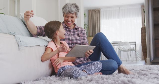 Happy caucasian grandmother and granddaughter sitting on floor, using tablet. Lifestyle, domestic life, communication, family, and togetherness.