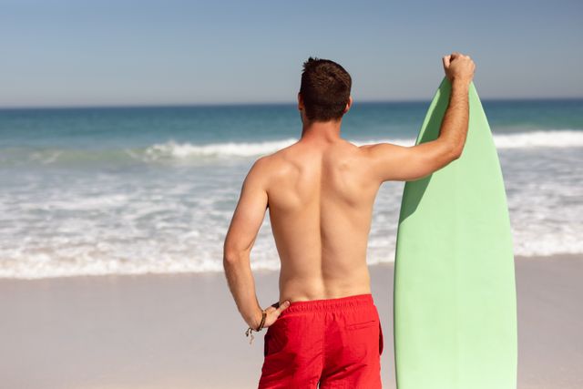 Rear view of shirtless man with surfboard standing on beach in the sunshine