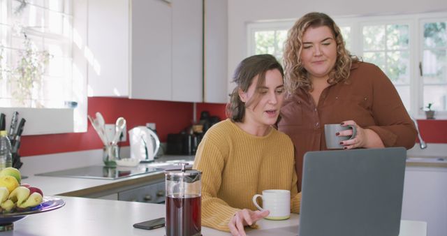 Happy caucasian lesbian drinking coffee and using laptop in kitchen. domestic life and leisure time.