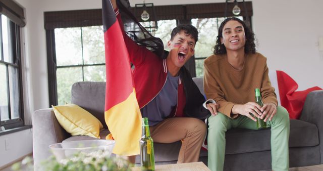 Happy diverse male couple watching tv with national flags and supporting. Spending quality time at home.