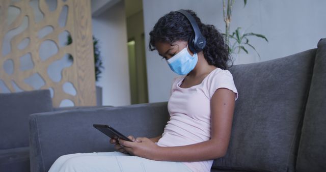 African american girl wearing face mask using digital tablet while sitting on the couch at hospital. medical healthcare during coronavirus covid 19 pandemic concept