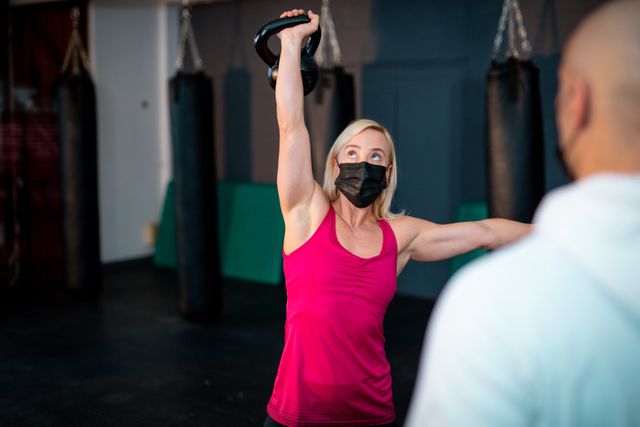 Caucasian woman wearing face mask lifting kettle ball on gym - Download Free Stock Photos Pikwizard.com