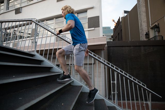 Side view of a fit Caucasian man with long blonde hair wearing sportswear exercising outdoors in the city on a sunny day with blue sky, running upstairs.