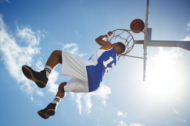 Low angle view of male teenager hanging on basketball hoop against blue sky