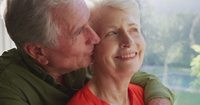 Happy, smiling senior caucasian couple smiling and embracing by window to garden. Love, togetherness, senior lifestyle and retirement.