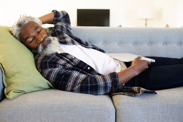 African American man enjoying time at home, social distancing and self isolation in quarantine lockdown, lying on sofa in sitting room, sleeping holding laptop.