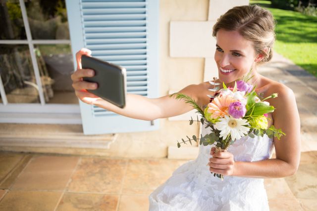 Happy bride holding bouquet taking selfie with mobile phone while sitting on chair in yard - Download Free Stock Photos Pikwizard.com