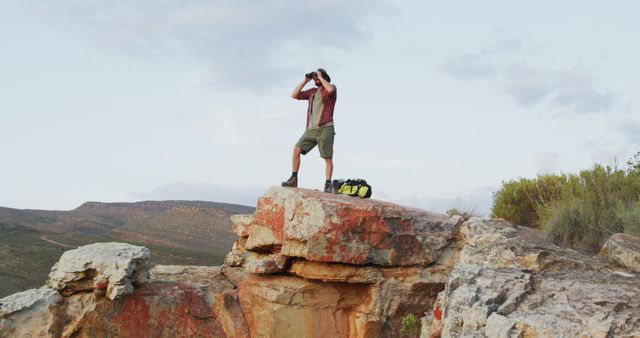 Caucasian male survivalist using binoculars, standing on mountain peak in wilderness. exploration, travel and adventure, survivalist in nature.