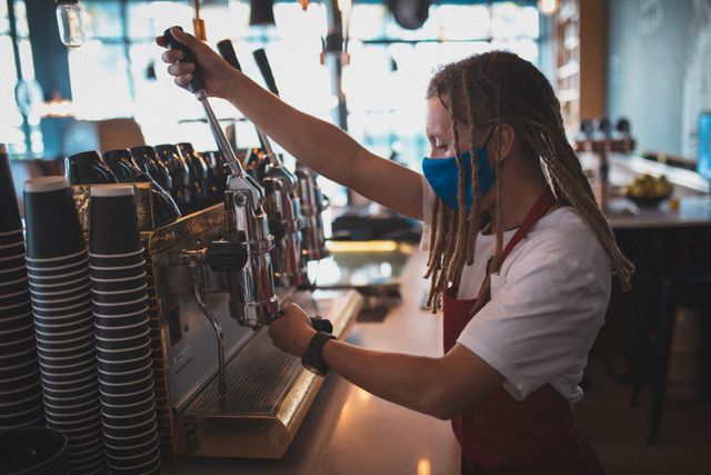 Mixed race male barista with dreadlocks wearing face mask and apron preparing coffee in cafe. independent small business during coronavirus covid 19 pandemic.