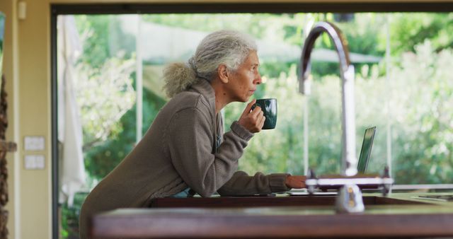 Portrait of senior mixed race woman holding mug and using laptop in kitchen. retirement and senior lifestyle, spending time alone at home.