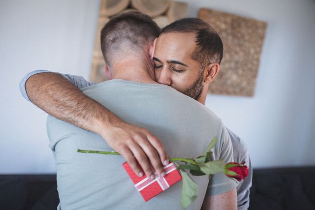 Diverse gay male hugging in living room one holding wrapped gift and red rose. staying at home in isolation during quarantine lockdown.