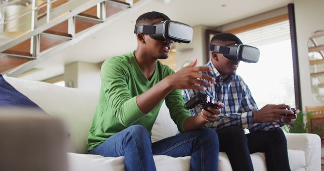African american teenage twin brothers sitting on couch using vr headsets and playing computer game. family leisure time at home together during quarantine lockdown.