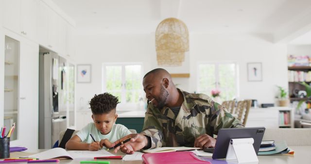 Happy african american male soldier and his son sitting at table, working and doing homework. Spending quality time together, army and patriotism concept.