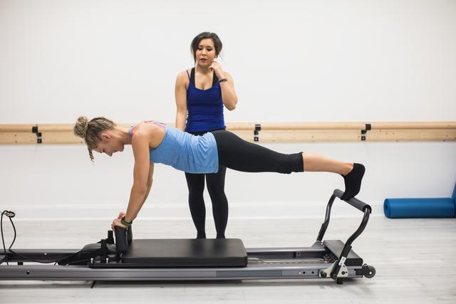 Female trainer assisting woman with stretching exercise on reformer - Download Free Stock Photos Pikwizard.com