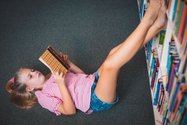 Relaxed caucasian schoolgirl lying on floor reading book in school library - Download Free Stock Photos Pikwizard.com