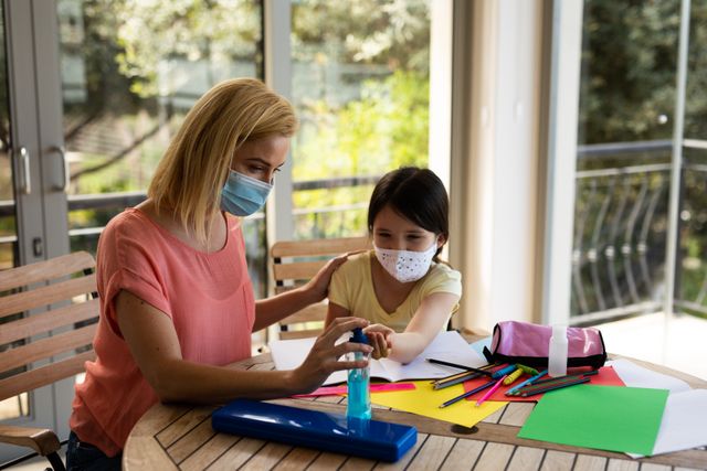 Caucasian mother squeezing out hand sanitizer for her daughter while they are studying at home. both of them are wearing masks.