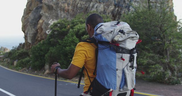 African american man wearing backpack trekking with walking poles on coastal road. Long distance walking, fitness, nature, and healthy outdoor lifestyle.