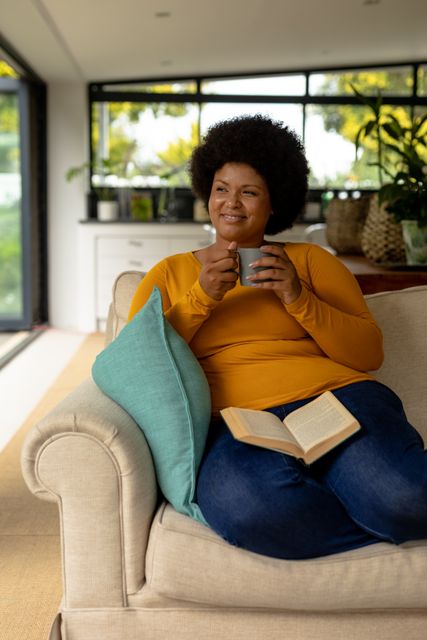 Smiling african american mid adult woman looking away while sitting with coffee cup and book. unaltered, lifestyle, leisure activity, drink, reading, hobbies and domestic life concept.